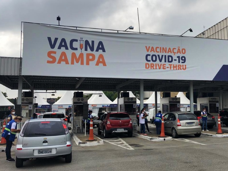 Foto do posto drive-thru do Anhembi. Acima há uma faixa com o texto Vacina Sampa - Vacinação Covid-19 Drive-Thru e alguns carros entrando pelas cabines. Agentes de saúde fazem o atendimento nos carros